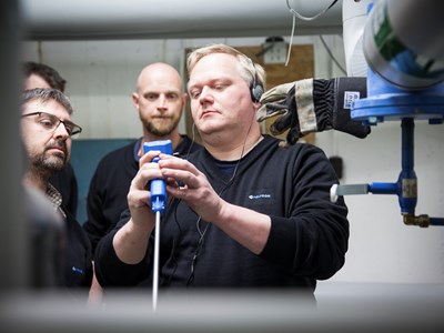 Tre tekniker i ett källarutrymme med rör i taket som studerar sin utrustning. 