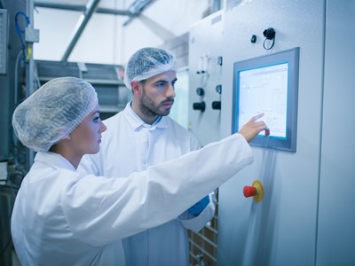 Två personer som står i vita skyddskläder och hårnät framför en skärm som är inbyggd i väggen. 